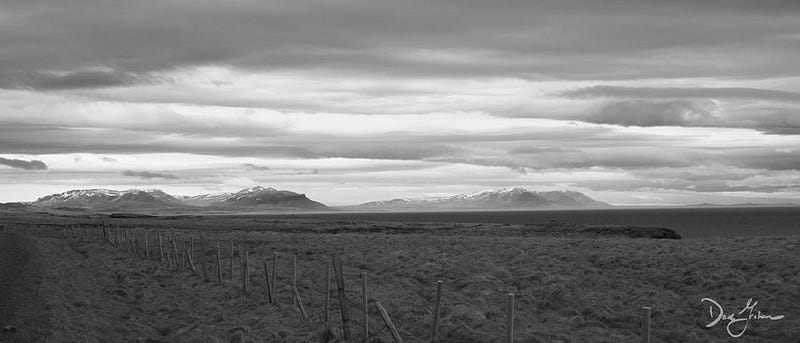 A cloudy gray sky over the mountains, ocean, and fields of Iceland, blockaded by a wood-and-wire fence.