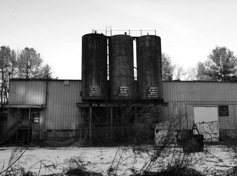 3 tall metal cylinders printed with “Do Not Enter” on their sides and attached to a broken-down metal warehouse with snow and